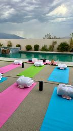 Poolside outdoor yoga setup with colorful pink, green, and blue mats, rolled white towels and props by a turquoise pool, desert plants and low wall with distant mountains under a dramatic stormy sky — ready for a wellness class.