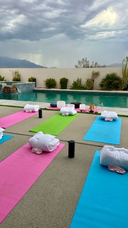 Poolside outdoor yoga setup with colorful pink, green, and blue mats, rolled white towels and props by a turquoise pool, desert plants and low wall with distant mountains under a dramatic stormy sky — ready for a wellness class.