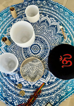 Top-down view of a blue-and-white mandala tapestry with crystal singing bowls, a frame drum, wooden mallet, tingsha cymbals and prayer beads arranged for a boho sound-healing meditation setup.