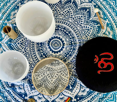 Top-down view of a blue-and-white mandala tapestry with crystal singing bowls, a frame drum, wooden mallet, tingsha cymbals and prayer beads arranged for a boho sound-healing meditation setup.