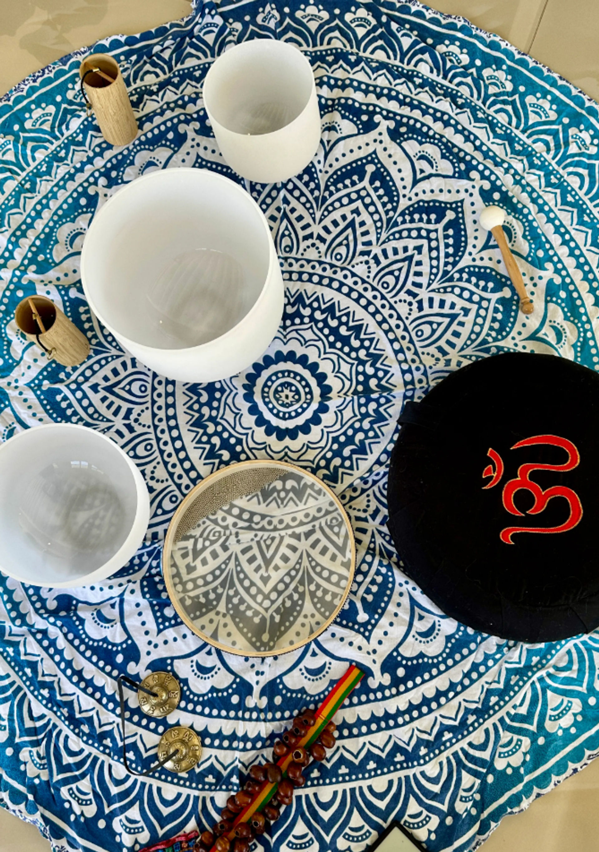 Top-down view of a blue-and-white mandala tapestry with white crystal singing bowls, a small tambourine, tingsha cymbals, wooden mallets, prayer beads and a black cushion embroidered with a red Om symbol—meditation sound-healing setup.