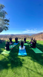 Outdoor yoga class on a grassy hilltop with colorful mats, participants in seated prayer pose overlooking a scenic lake and rolling hills under a bright blue sky