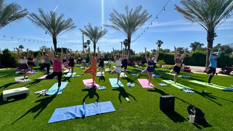 Outdoor group yoga class on a sunny palm-lined lawn, participants holding tree pose on colorful mats under a bright blue sky with string lights overhead.