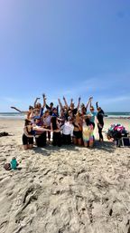 Large group of friends cheering and posing in the sun on a sandy beach with ocean waves in the background, colorful towels and a cooler at the right — playful summer beach group photo.