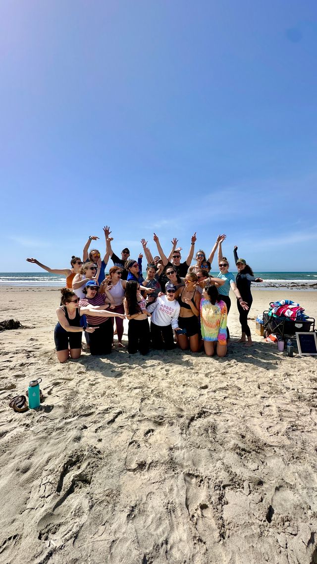 Large group of friends cheering and posing in the sun on a sandy beach with ocean waves in the background, colorful towels and a cooler at the right — playful summer beach group photo.