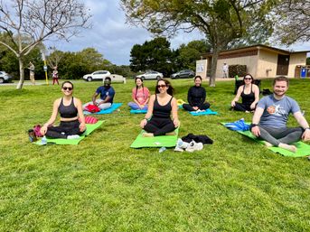 Smiling adults in workout clothes seated on bright yoga mats during an outdoor yoga class on a sunny green park lawn with trees, parked cars and a restroom building in the background.