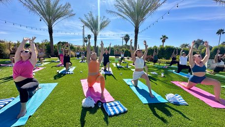 Sun-soaked outdoor group yoga class on a green lawn with palm trees and string lights, participants on colorful mats in a lunge pose reaching upward under a bright blue sky