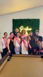 Cheerful indoor bachelorette party — ten women wearing pink sashes posing in front of a green boxwood wall with a neon "Sunshine State" sign and a pool table in the foreground.