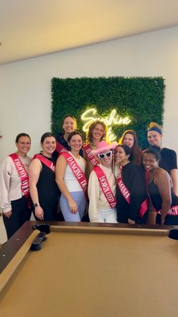 Cheerful indoor bachelorette party — ten women wearing pink sashes posing in front of a green boxwood wall with a neon "Sunshine State" sign and a pool table in the foreground.