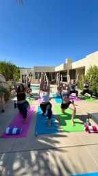 Poolside outdoor yoga class on colorful mats in a sunny modern backyard patio under a clear blue sky, participants in high lunge with arms raised.
