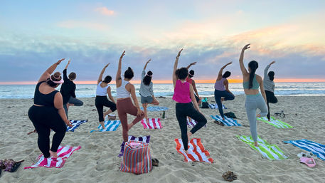 Sunset beach yoga class: a group doing tree pose on striped towels on a sandy shore with the ocean and pastel sky in the background.