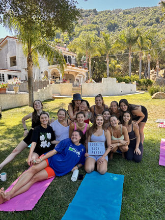 Group of smiling women posing on yoga mats for a bachelorette bach party on a sunny Southern California hillside lawn in front of a Mediterranean-style villa with palm trees.