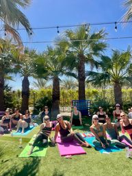 Group of women in athletic wear smiling on colorful yoga mats during an outdoor yoga session on a sunny palm-tree lawn with string lights overhead