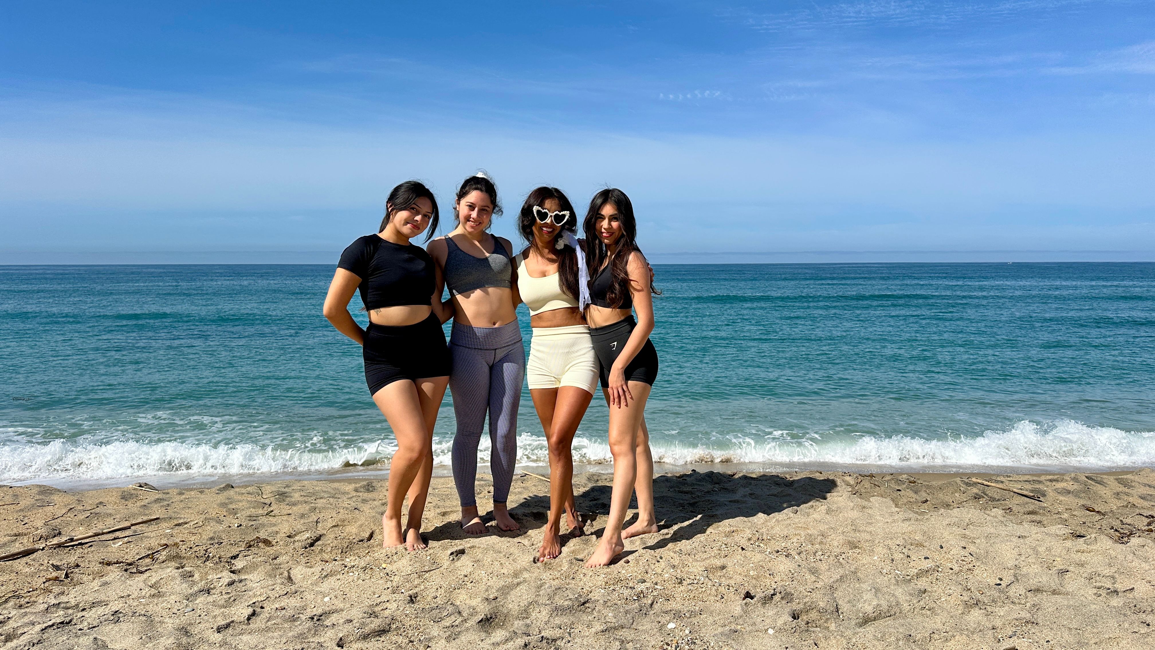 Four women in activewear barefoot on a sunny sandy beach, posing by turquoise ocean waves under a clear blue sky