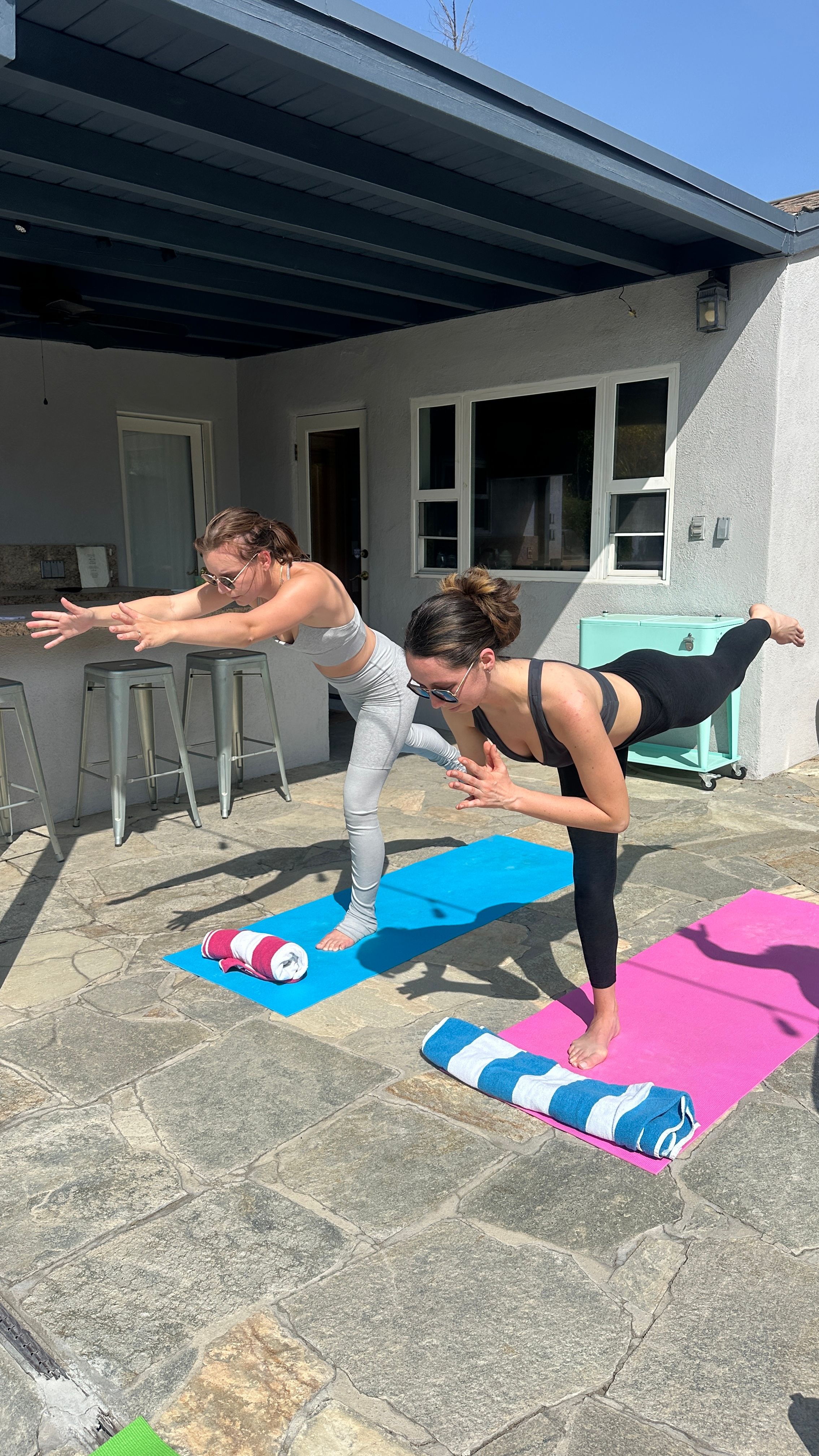 Two women practicing outdoor yoga balancing poses on pink and blue mats on a sunny stone backyard patio under a covered porch, with rolled towels and metal barstools nearby.