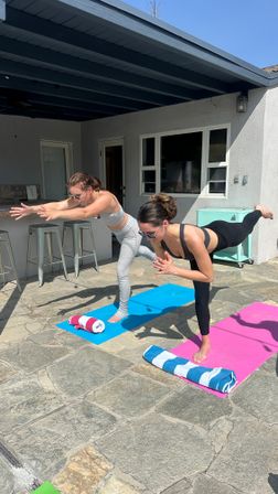 Two women practicing outdoor yoga balancing poses on pink and blue mats on a sunny stone backyard patio under a covered porch, with rolled towels and metal barstools nearby.