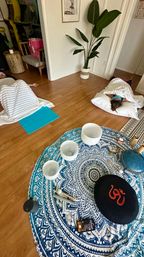 Cozy indoor meditation scene in a home yoga studio: blue mandala rug with crystal singing bowls, mallets, Om cushion and handpan nearby, while two people rest on pillows under white blankets on a hardwood floor.