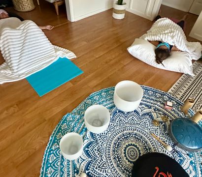 At-home living room sound bath — two people lying on hardwood floor under white blankets with pillows, nearby blue mandala mat holding crystal singing bowls, mallets and a metal tongue drum, potted plant in the corner.