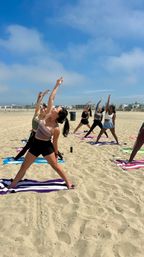 Group beach yoga: women in athletic wear stretching in standing side poses on towels on a sunny sandy shore under a bright blue sky with beachfront buildings in the distance.
