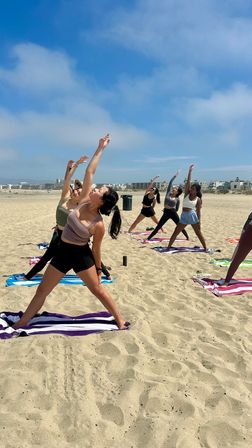 Group beach yoga: women in athletic wear stretching in standing side poses on towels on a sunny sandy shore under a bright blue sky with beachfront buildings in the distance.