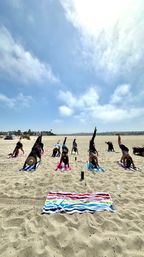Group beach yoga class on sunny coastal sand, participants on colorful towels and mats holding downward-dog split poses beneath a bright blue sky.