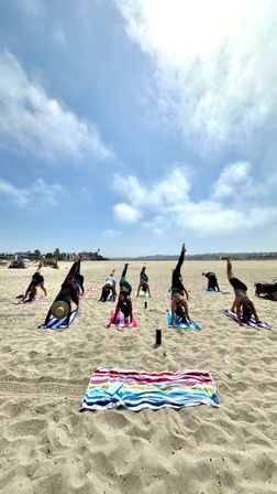 Group beach yoga class on sunny coastal sand, participants on colorful towels and mats holding downward-dog split poses beneath a bright blue sky.