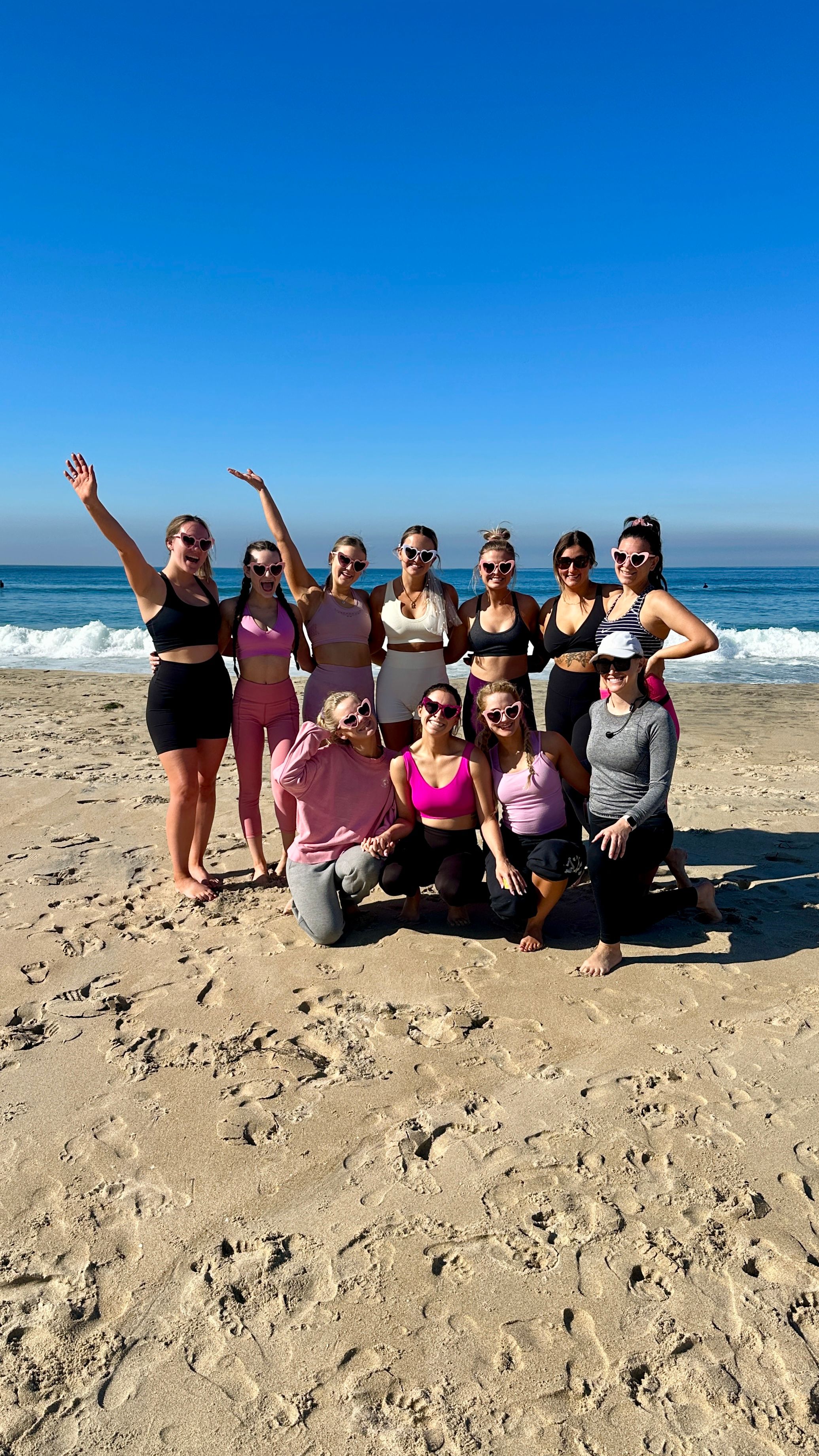 Group of women in colorful activewear and heart-shaped sunglasses posing on a sunny sandy oceanfront beach with gentle waves and a clear blue sky