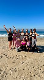 Group of women in colorful activewear and heart-shaped sunglasses posing on a sunny sandy oceanfront beach with gentle waves and a clear blue sky