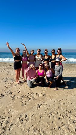 Group of women in colorful activewear and heart-shaped sunglasses posing on a sunny sandy oceanfront beach with gentle waves and a clear blue sky