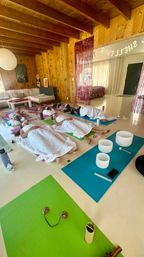 Cozy wood‑paneled retreat room hosting a group sound‑bath meditation — participants reclined on yoga mats under white blankets with crystal singing bowls, cymbals, water bottles, and soothing decor visible.