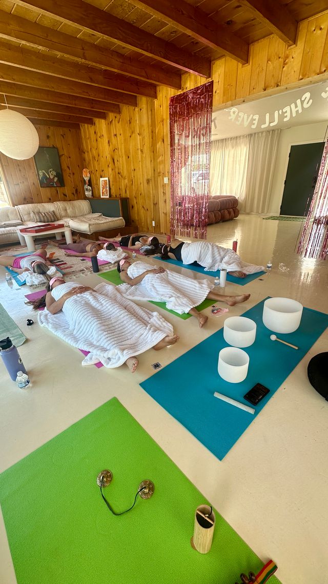 Restorative sound-bath session in a cozy wood-paneled wellness studio — participants lying on colorful yoga mats under white blankets, crystal singing bowls and mallet on a blue mat, small cymbals, water bottles and a pink fringe curtain nearby.