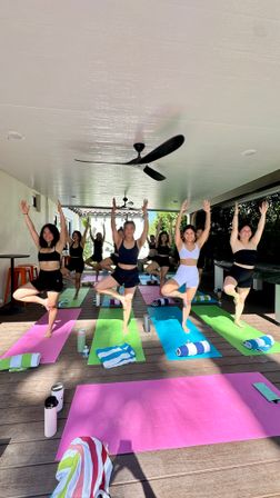 Sunlit group yoga class on a covered outdoor deck, participants balancing in tree pose on colorful mats with towels and water bottles, ceiling fans overhead and lush greenery beyond.