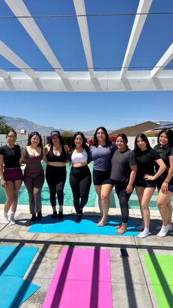 Group of eight women in activewear posing arm-in-arm poolside under a white pergola with colorful yoga mats, turquoise pool water and mountain backdrop under a clear blue sky