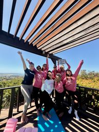 Group of eight women in matching pink shirts celebrating a bachelorette on a sunny San Diego deck under a wooden pergola, posing playfully on a blue yoga mat with arms raised and coastal view in the background.