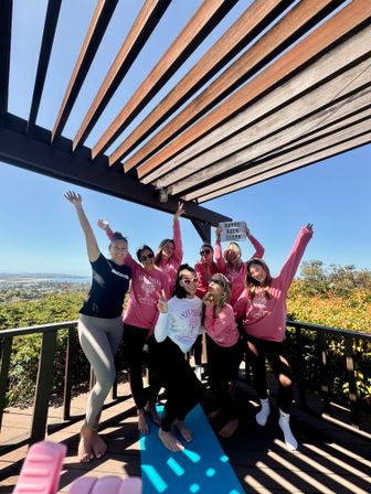Group of eight women in matching pink shirts celebrating a bachelorette on a sunny San Diego deck under a wooden pergola, posing playfully on a blue yoga mat with arms raised and coastal view in the background.