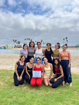 Ten friends in colorful activewear posing on a grassy beach by a lagoon with palm trees and a cloudy sky, center holds a lightbox sign reading "HAPPY BACH SUNMEET" — bachelorette beach vibe