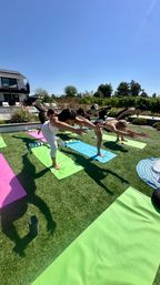 Outdoor yoga class on a sunny blue-sky day — a group of people balancing in one-legged forward poses on colorful mats on a green backyard lawn with a modern house and trees in the background.