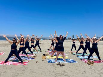 Group of women in black activewear doing star-stretch poses on colorful beach towels during a sunny beach fitness class with sand, ocean and clear blue sky in the background