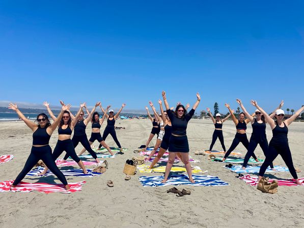 Group of women in black activewear doing star-stretch poses on colorful beach towels during a sunny beach fitness class with sand, ocean and clear blue sky in the background