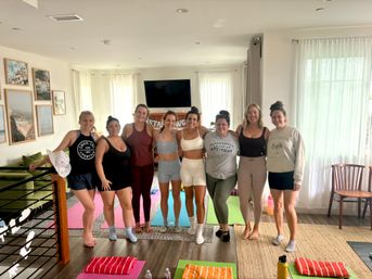 Eight women in workout gear posing on colorful yoga mats in a sunlit living-room home studio after a group yoga/fitness session, casual at-home wellness meetup.
