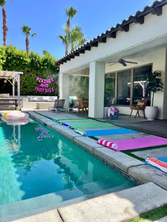 Sunny Palm Springs–style backyard pool with tall palm trees, colorful mats and striped towels lined along the deck, pastel inflatable float, covered patio, and neon "BIKINIS AND MARTINIS" sign