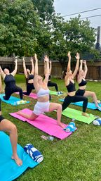 Outdoor backyard yoga class with women in high lunge pose, arms raised on colorful mats on a green lawn beneath trees and a wooden fence, towels and water bottles nearby.