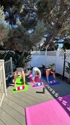Three people holding plank pose on colorful yoga mats (green, pink, blue) in a sunny backyard deck yoga session under a large pine tree, white lattice fence, with rolled towels and water bottles nearby.