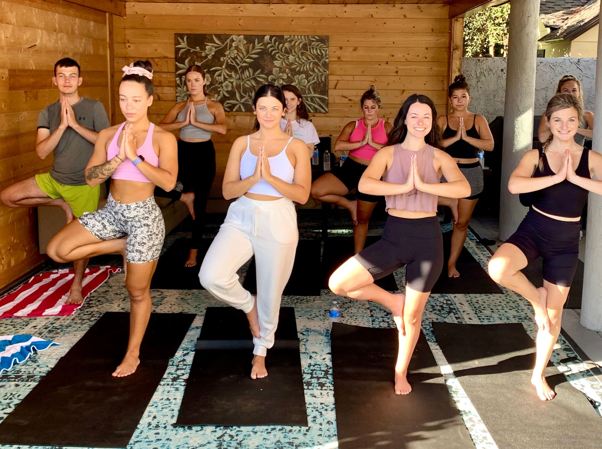Group yoga class holding tree pose on mats in a sunlit wood-paneled outdoor studio/patio, participants in athletic wear with palms pressed together — lively group fitness/wellness session.