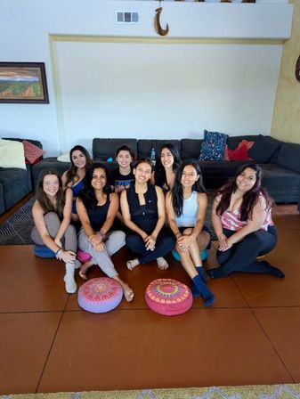 Smiling group of nine women seated on a living room floor in a semicircle with colorful round meditation cushions in front and a dark sectional sofa with decorative pillows behind, casual wellness gathering vibe.