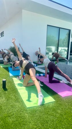 Sunny outdoor yoga class with several people doing side-plank on colorful mats on artificial turf beside a modern white house and pool