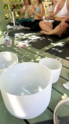 Outdoor yoga and sound bath on a shaded wooden deck — white crystal singing bowls in the foreground, participants seated cross‑legged on black mats with hands in prayer pose under dappled sunlight.