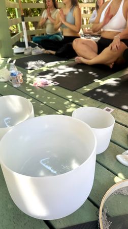 Outdoor yoga and sound bath on a shaded wooden deck — white crystal singing bowls in the foreground, participants seated cross‑legged on black mats with hands in prayer pose under dappled sunlight.