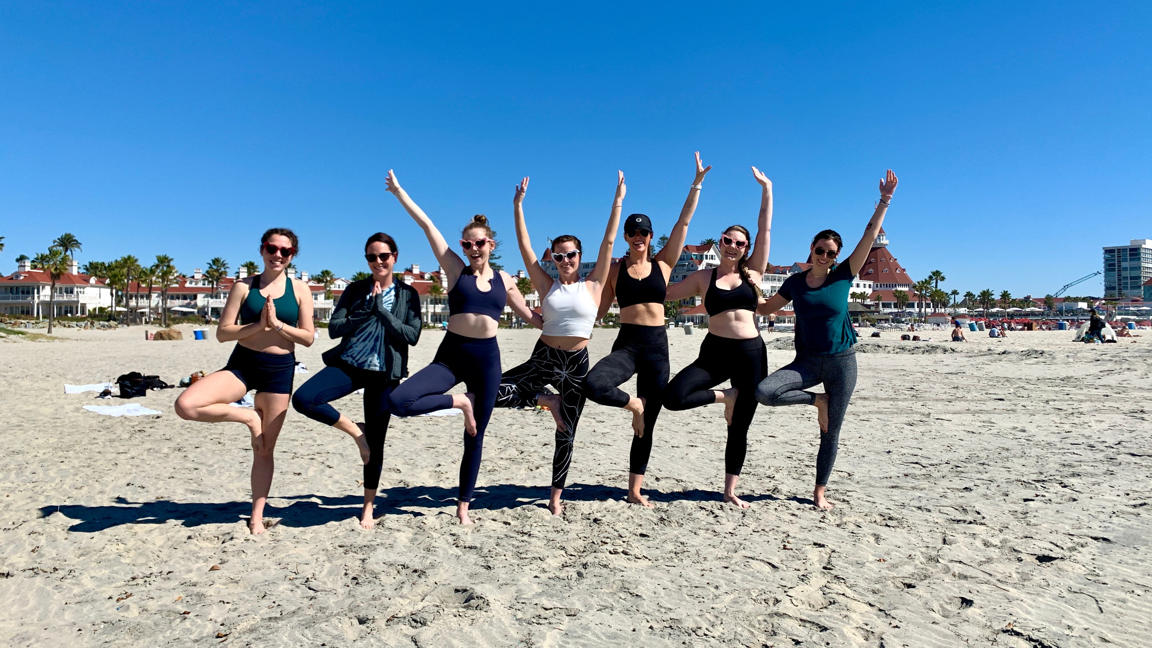 Group of seven women in activewear doing tree-pose yoga in a line on a sunny sandy beach with palm trees and oceanfront buildings under a clear blue sky