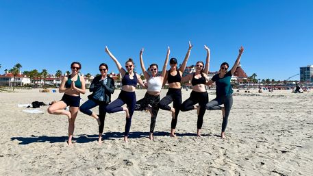 Group of seven women in activewear doing tree-pose yoga in a line on a sunny sandy beach with palm trees and oceanfront buildings under a clear blue sky